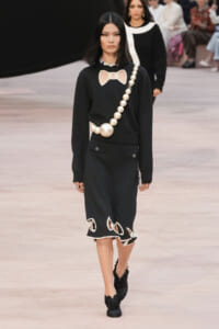Model walks runway in black knit outfit with oversized pearl necklace and cutout bow detail on chest, black skirt with scalloped white trim, and black footwear.