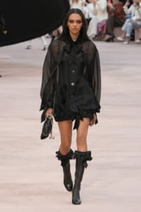 Model walks the runway in a sheer black blouse with a large bow at the neck, a black mini dress, and knee-high black boots with bows, carrying a small black handbag.