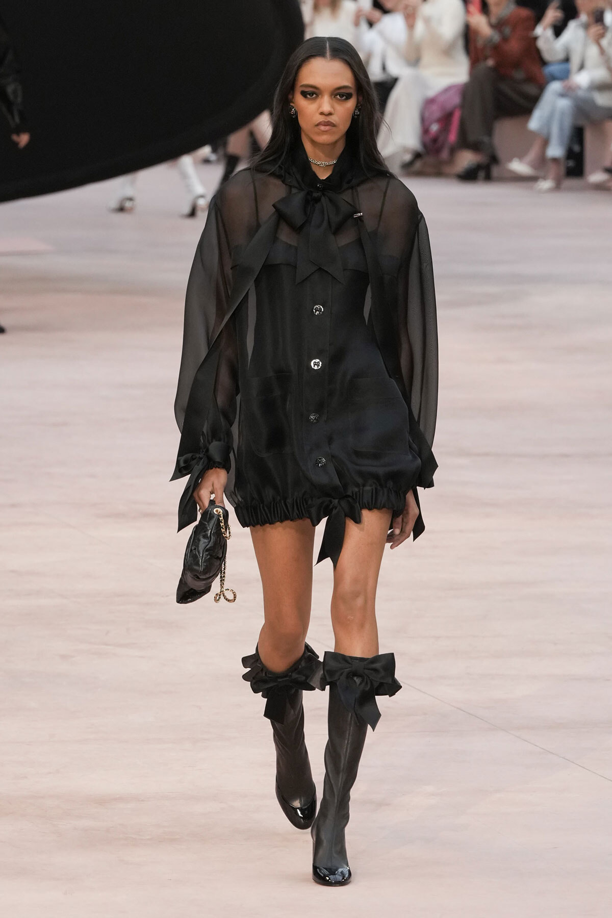 Model walks the runway in a sheer black blouse with a large bow at the neck, a black mini dress, and knee-high black boots with bows, carrying a small black handbag.