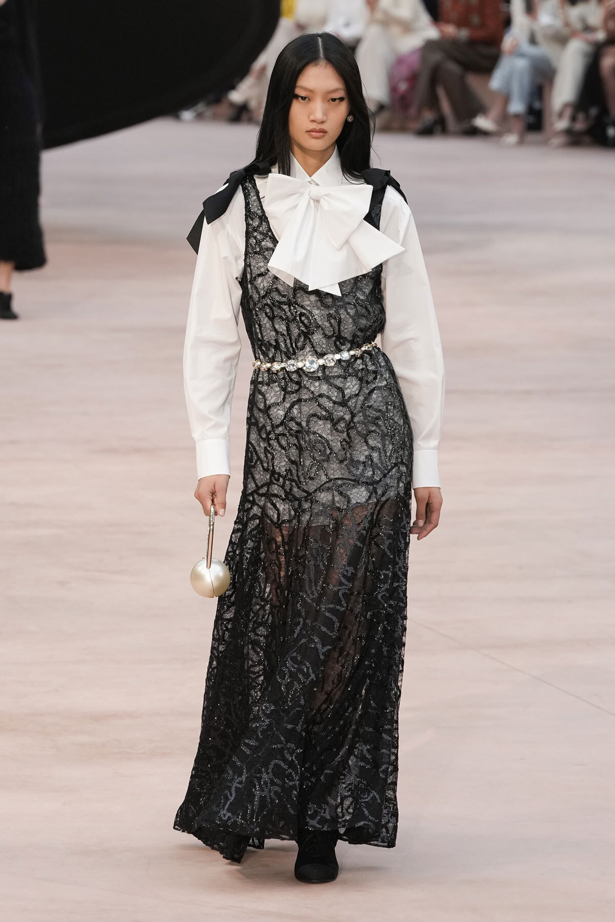 Model on a runway wearing a white shirt with a large bow, layered under a black lace gown, with a pearl belt and a round metallic handbag.