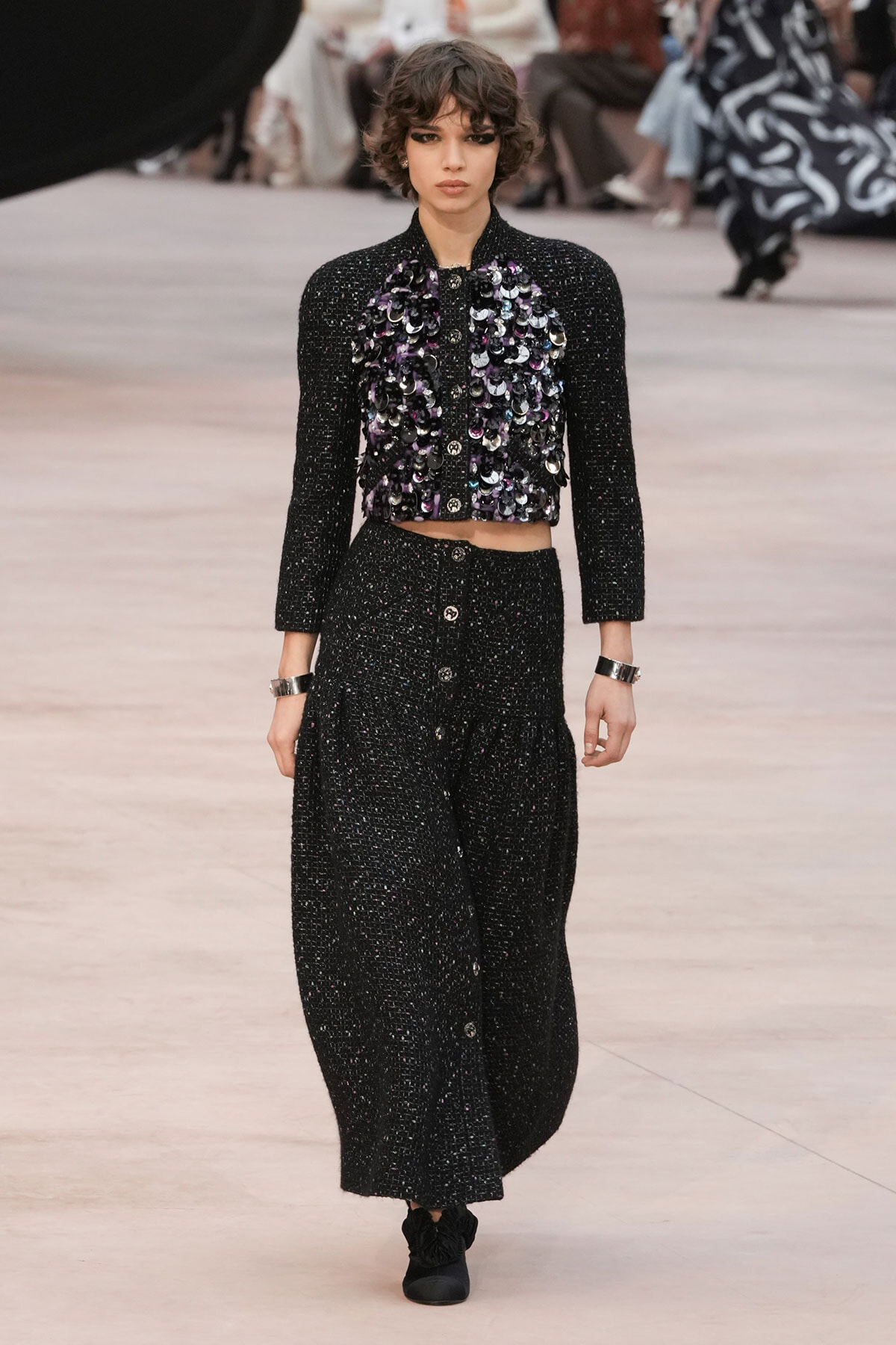 Model walking the runway in a black sequined cropped jacket with a purple-iris sequin panel, paired with matching high-waisted wide-leg trousers and black shoes.
