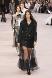 Model walks the runway in a black tweed double-breasted coat with a sheer tulle overlay and chunky jewelry, centered with audience blurred in the background.