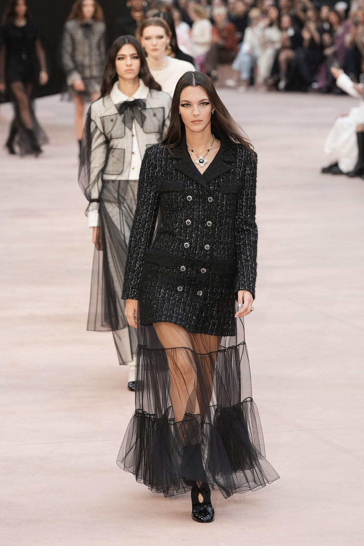 Model walks the runway in a black tweed double-breasted coat with a sheer tulle overlay and chunky jewelry, centered with audience blurred in the background.