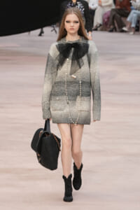 Model walking a runway in a grey gradient knit mini dress with a large black tulle bow at the neck and pearl accessories, carrying a black handbag and wearing black ankle boots.