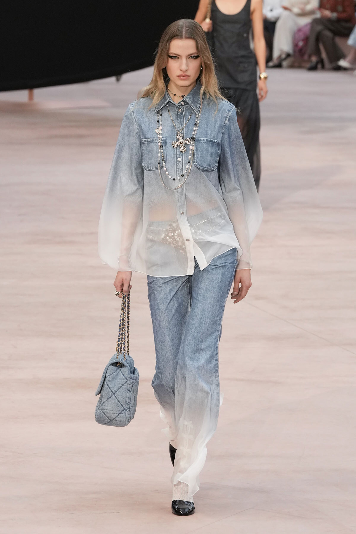 Model on a runway wearing an ombré denim shirt with matching jeans and layered pearl necklaces, carrying a quilted blue handbag on a chain strap.