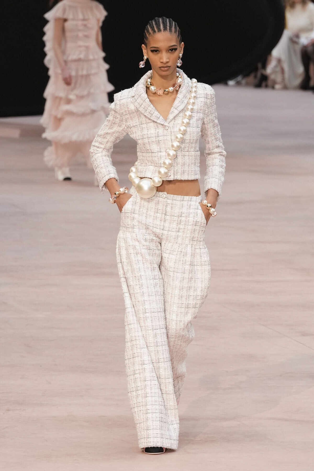 Model walks the runway in a white tweed suit with matching blazer and wide-leg pants, accessorized with oversized pearl jewelry and earrings.