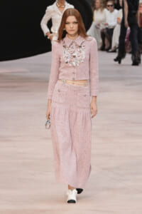 Model wearing a pink tweed cropped jacket and wide-leg trousers with a heavy silver necklace, walking a runway.