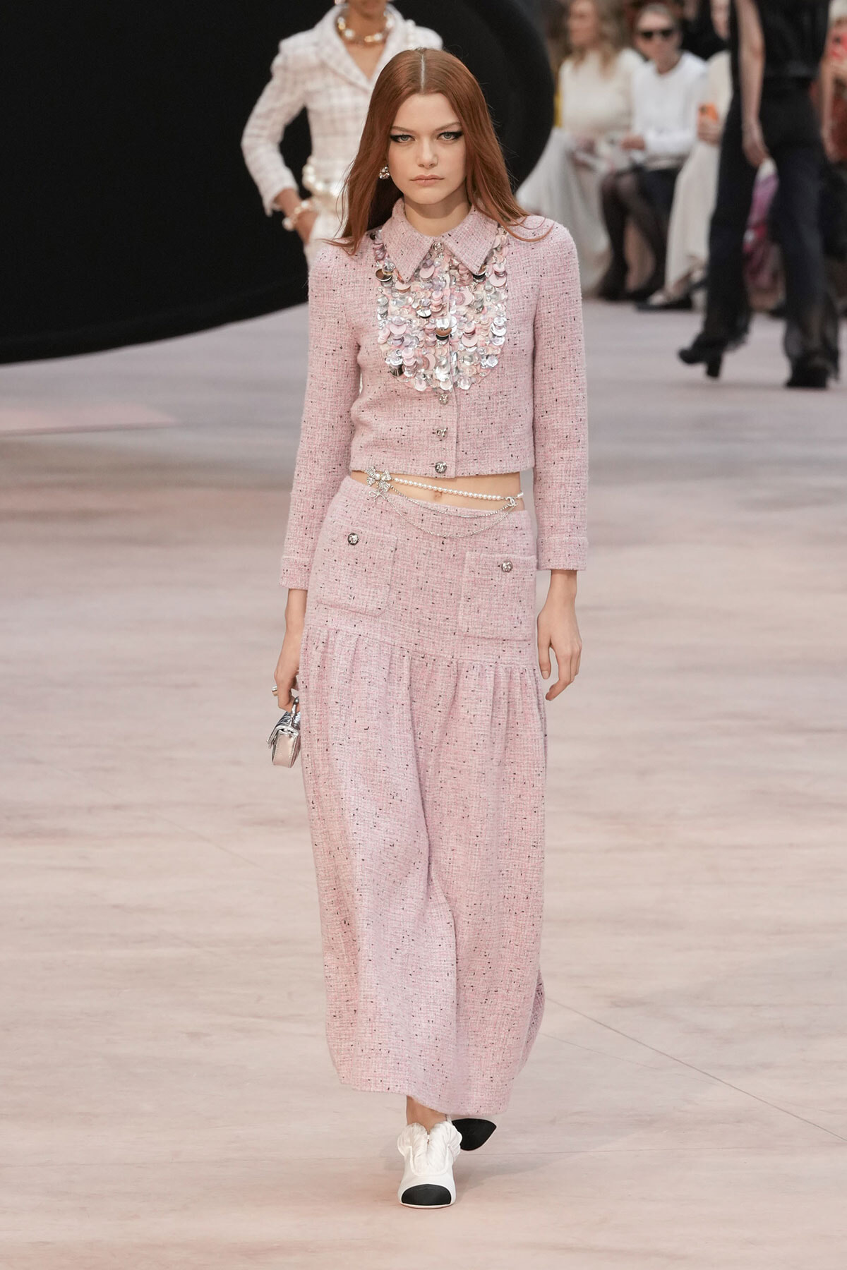 Model wearing a pink tweed cropped jacket and wide-leg trousers with a heavy silver necklace, walking a runway.