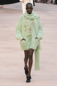 Model on a runway wearing a pale mint chunky knit oversized cardigan with a matching knit scarf and long pearl necklaces; black shoes with ruffle accents.