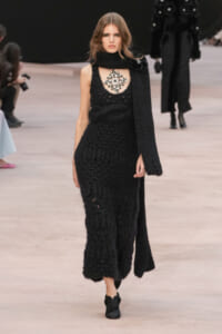 Model on a runway wearing a long black knit dress with a matching scarf and a large jeweled necklace, black shoes.