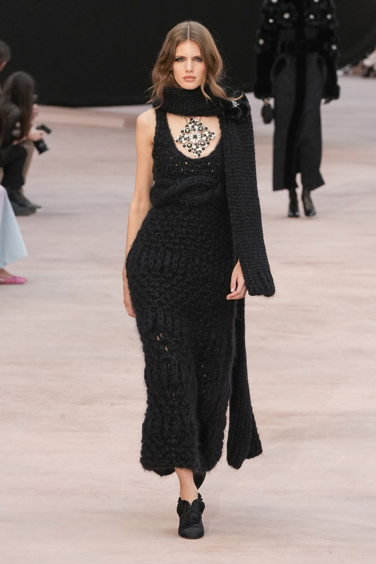 Model on a runway wearing a long black knit dress with a matching scarf and a large jeweled necklace, black shoes.