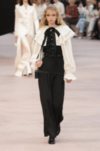 Model walking the runway in a white ruffled blouse with black bow and black patterned wide-leg pants, accessorized with statement earrings and a small handbag.