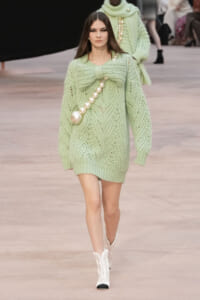 Model walks down a runway in a pale mint-green oversized knit sweater dress with a large bow at the chest and a pearl bead strap, paired with white boots.
