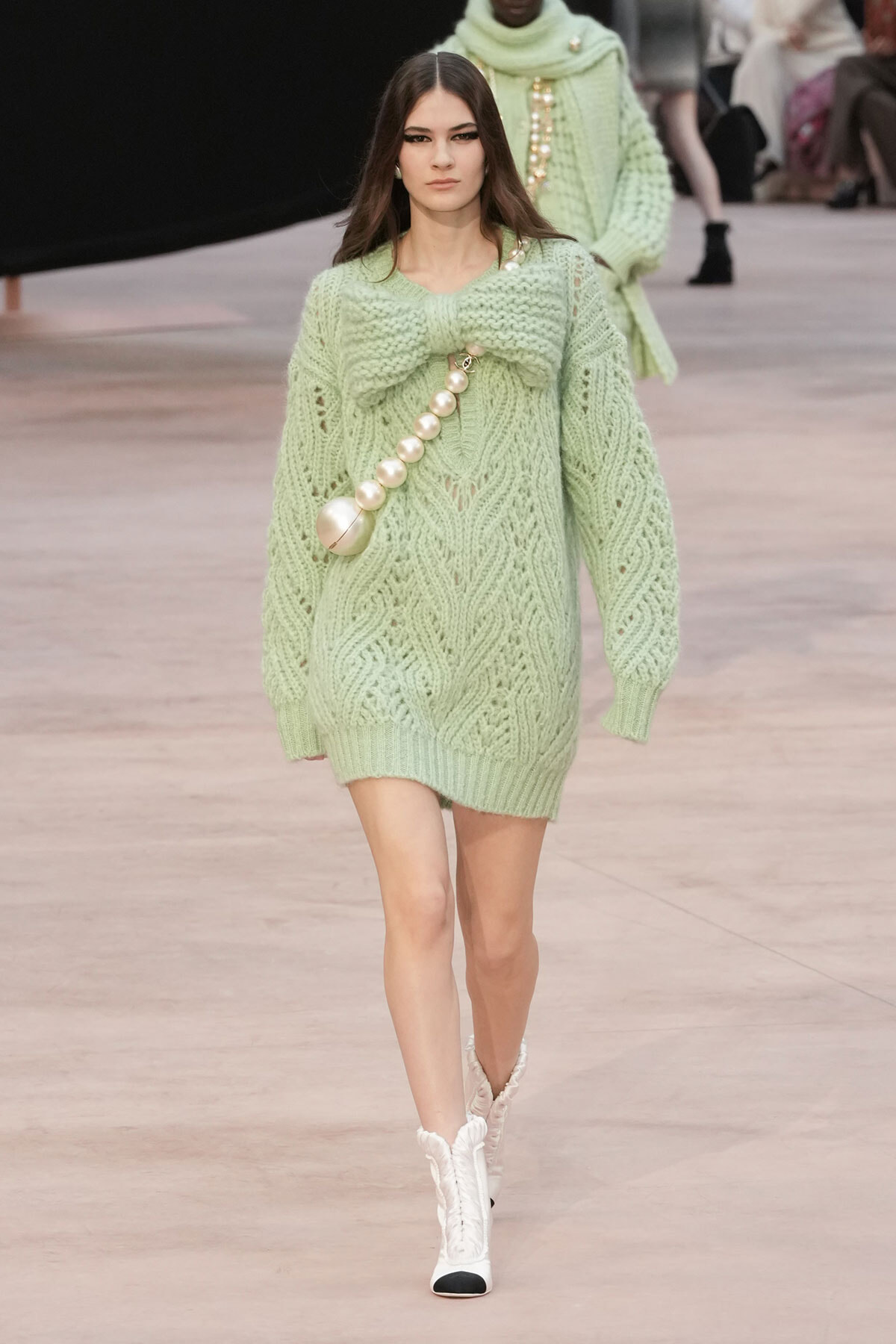 Model walks down a runway in a pale mint-green oversized knit sweater dress with a large bow at the chest and a pearl bead strap, paired with white boots.