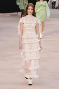 Model walking down a runway in a pale pink, tiered ruffle gown with bow accents and a cape-like overlay, red hair and statement earrings visible.