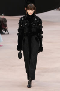 Female model walking a runway in an all-black fur coat with large velvet flower appliqués and jewel embellishments, holding a small matching purse.