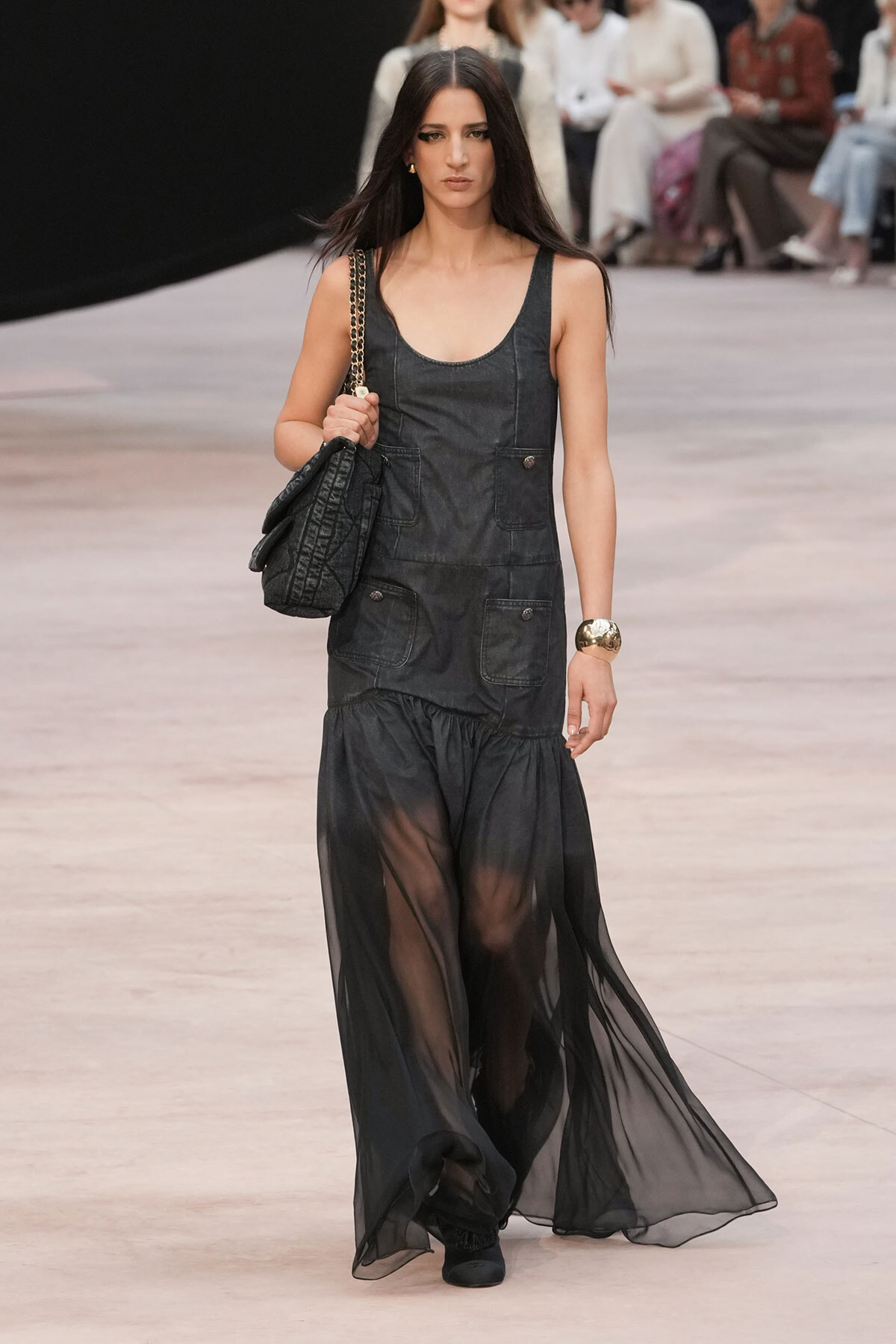 Model walking the runway in a sleeveless dark denim jumpsuit with patch pockets, a sheer black skirt overlay, black shoes, a gold cuff bracelet, and a quilted chain-strap bag.