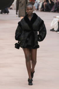 Model walking the runway in a black fur-coated ensemble with a small black handbag and braided hair.