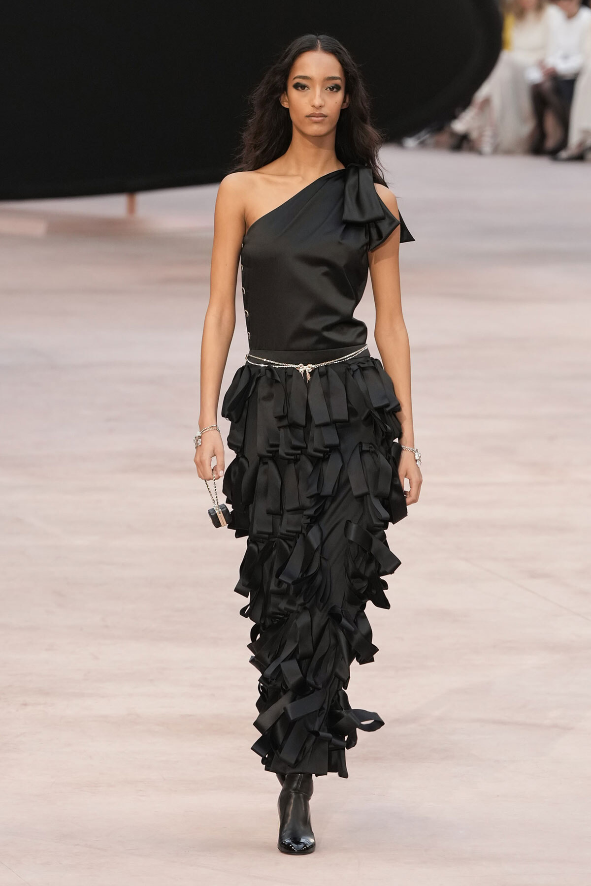 Female model walking a fashion runway in a black one-shoulder satin top and multi-tiered ruffle skirt with a sparkling belt and ankle boots.