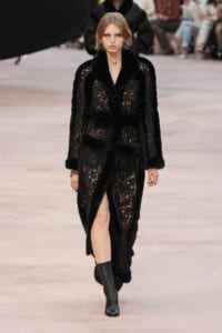 Model walking a fashion runway in a black lace fur-trim coat with a wide belt and thigh-high slit, paired with black ankle boots.