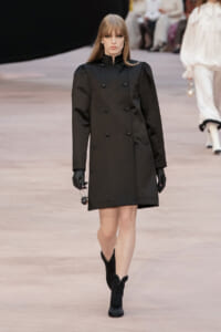 Model walking a runway in a black double-breasted coat, black gloves, and black heeled boots, holding a small round handbag.