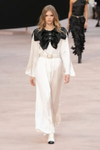 Model walks the runway in a white, wide-leg jumpsuit with a dramatic black bow neckline and pearl belt.
