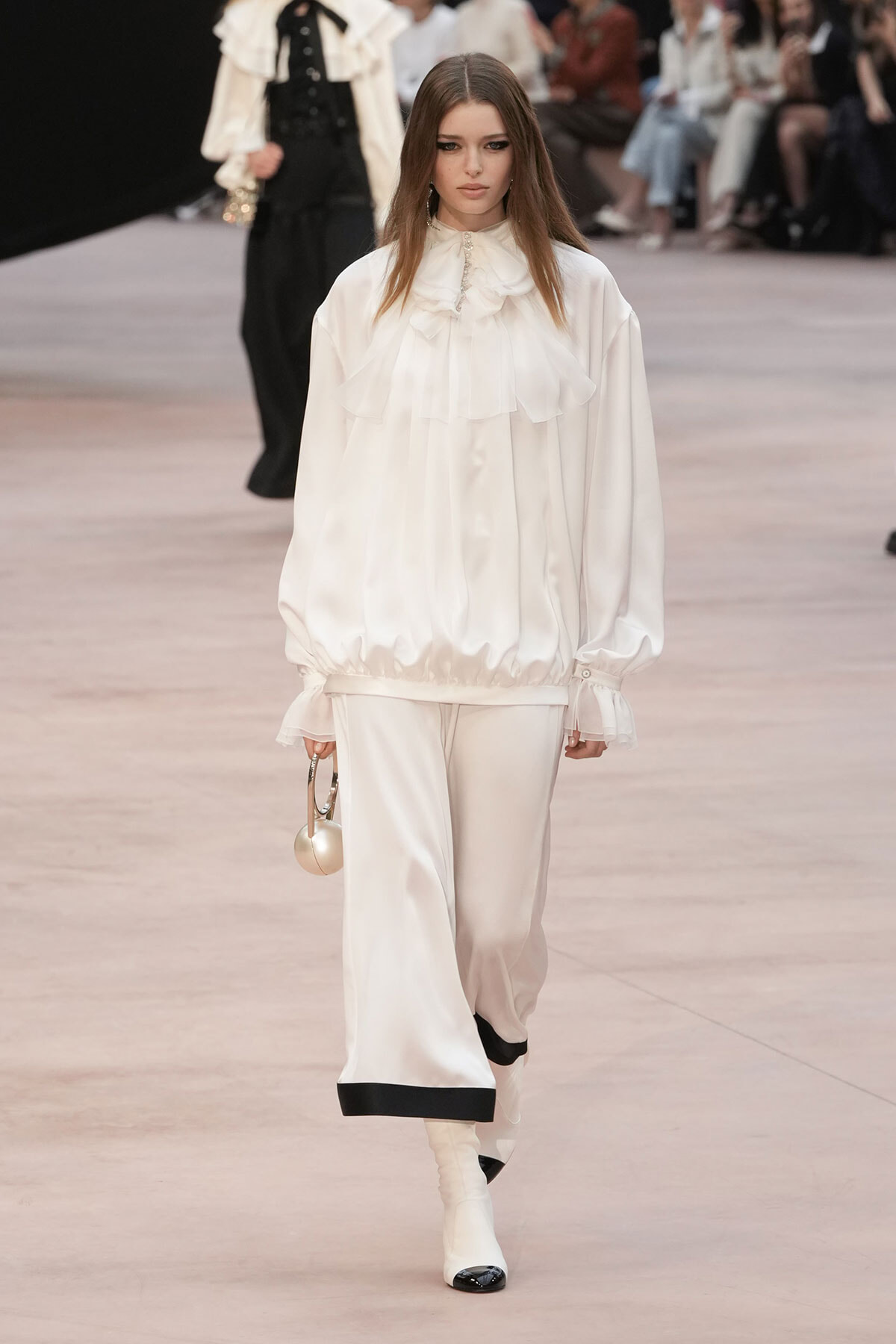 Model walking a runway in an all-white satin outfit with a ruffled blouse and wide-leg pants, carrying a small round handbag on the left.