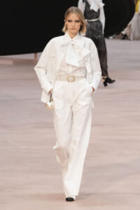 Model walking the runway in an all-white suit with oversized bow blouse and pearl belt, holding a round metallic bag.