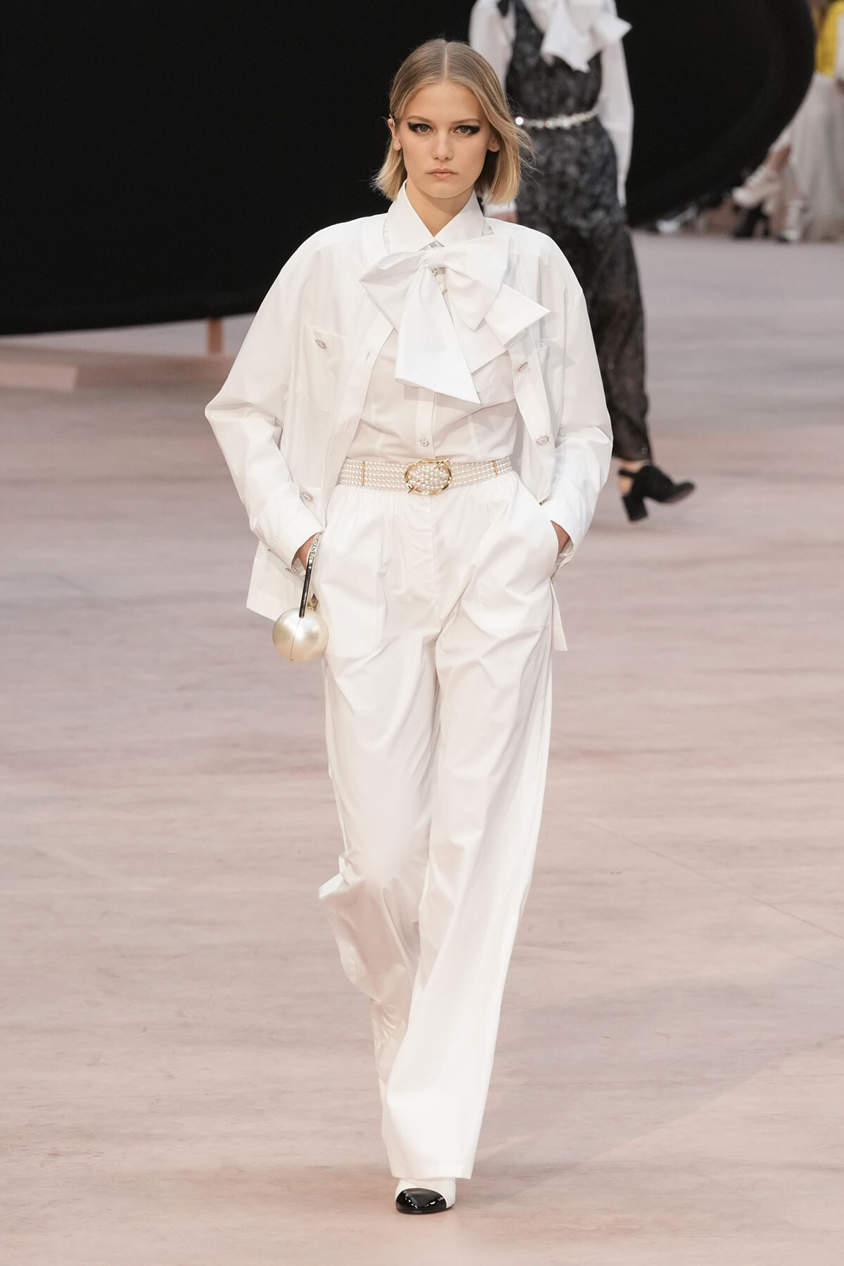 Model walking the runway in an all-white suit with oversized bow blouse and pearl belt, holding a round metallic bag.