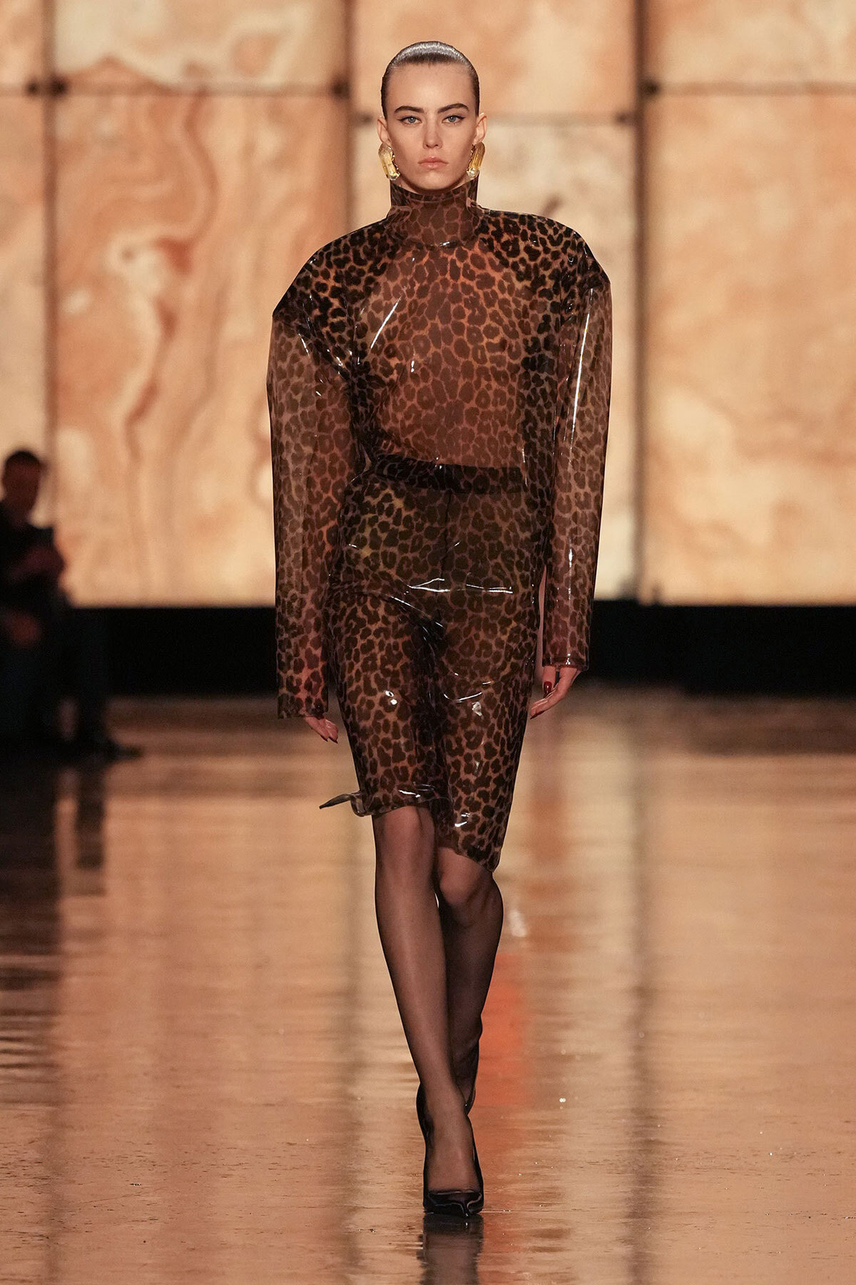 Model walks a fashion show in a sheer leopard-print dress with a high neck and long sleeves, paired with black tights and heels.