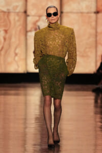Model on the runway wearing a gold lace high-neck blouse and a green lace knee-length skirt, with black heels and oversized sunglasses.