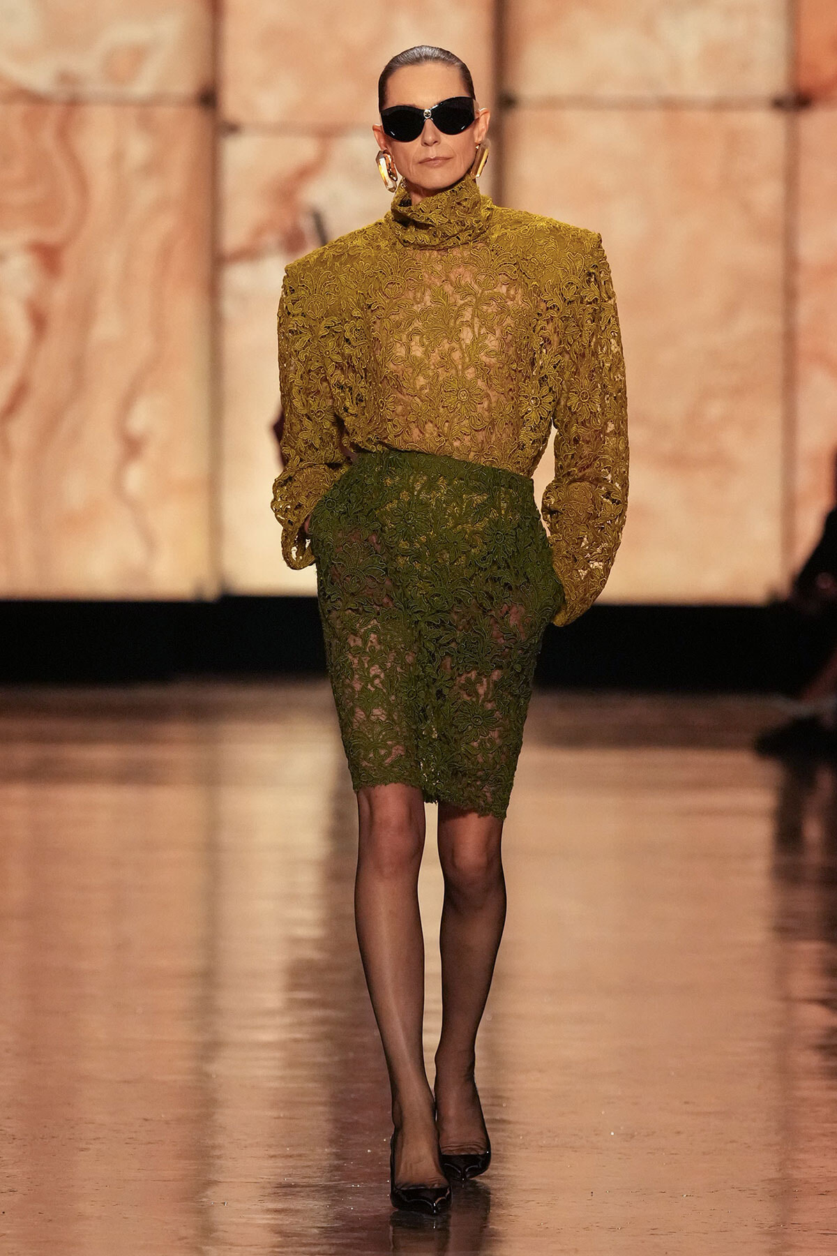 Model on the runway wearing a gold lace high-neck blouse and a green lace knee-length skirt, with black heels and oversized sunglasses.