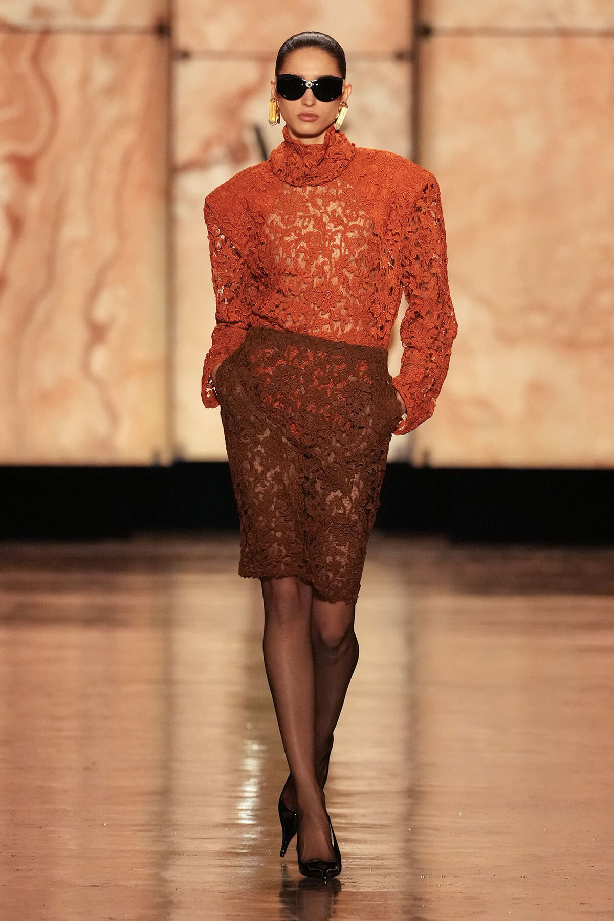Model on a runway wearing a burnt-orange lace blouse with high neck and a brown lace pencil skirt, black tights, heels, and oversized sunglasses.