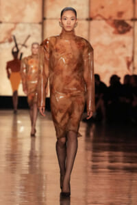 Runway model in a translucent brown floral dress with a high neck and long sleeves, paired with black tights and heels.