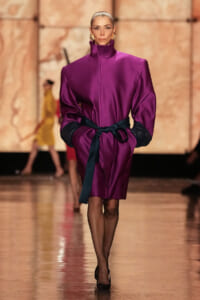 Model walking the runway in a magenta satin belted coat with oversized sleeves and black gloves, gold earrings, and black heels in a warm-toned backdrop.