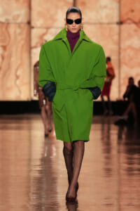 Model on a runway wearing a bright green oversized coat tied at the waist, black gloves, purple turtleneck and large sunglasses with gold earrings.
