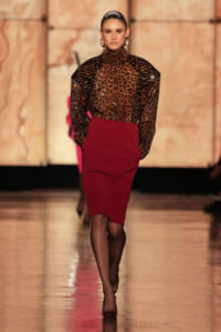 Model on a runway wearing a sheer leopard-print blouse tucked into a burgundy pencil skirt, with gold earrings and black tights.