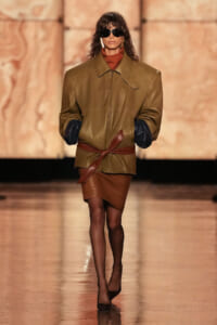 Model wearing oversized tan leather jacket with dark cuffed sleeves, tied at the waist with a brown belt, on a fashion runway.