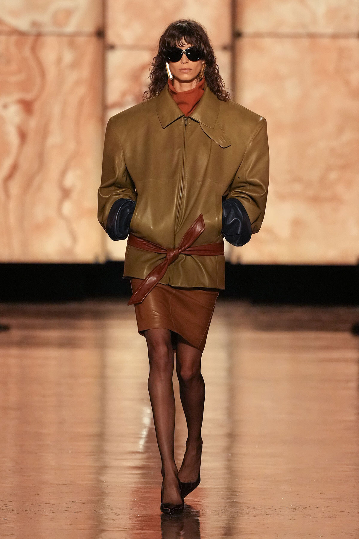 Model wearing oversized tan leather jacket with dark cuffed sleeves, tied at the waist with a brown belt, on a fashion runway.