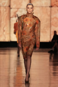 Model walks runway wearing a sheer, brown-toned floral dress with a turtleneck; the translucent fabric reveals a matching underlayer as she poses in a fashion show studio.