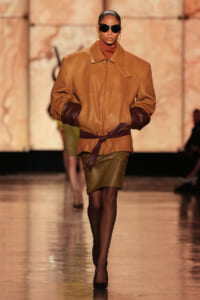 Model walks a runway wearing a camel tan oversized leather jacket with dark brown cuffed sleeves, a matching belt, and a knee-length olive skirt.