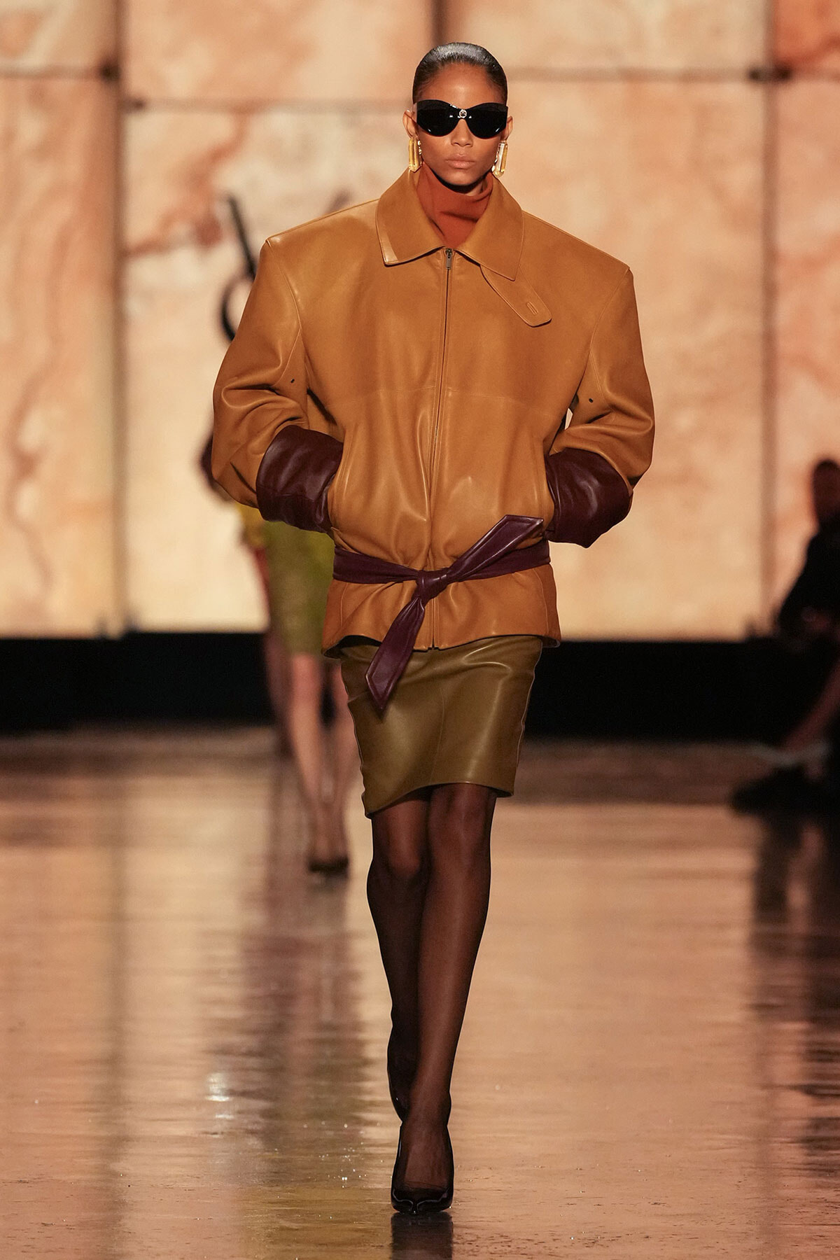 Model walks a runway wearing a camel tan oversized leather jacket with dark brown cuffed sleeves, a matching belt, and a knee-length olive skirt.