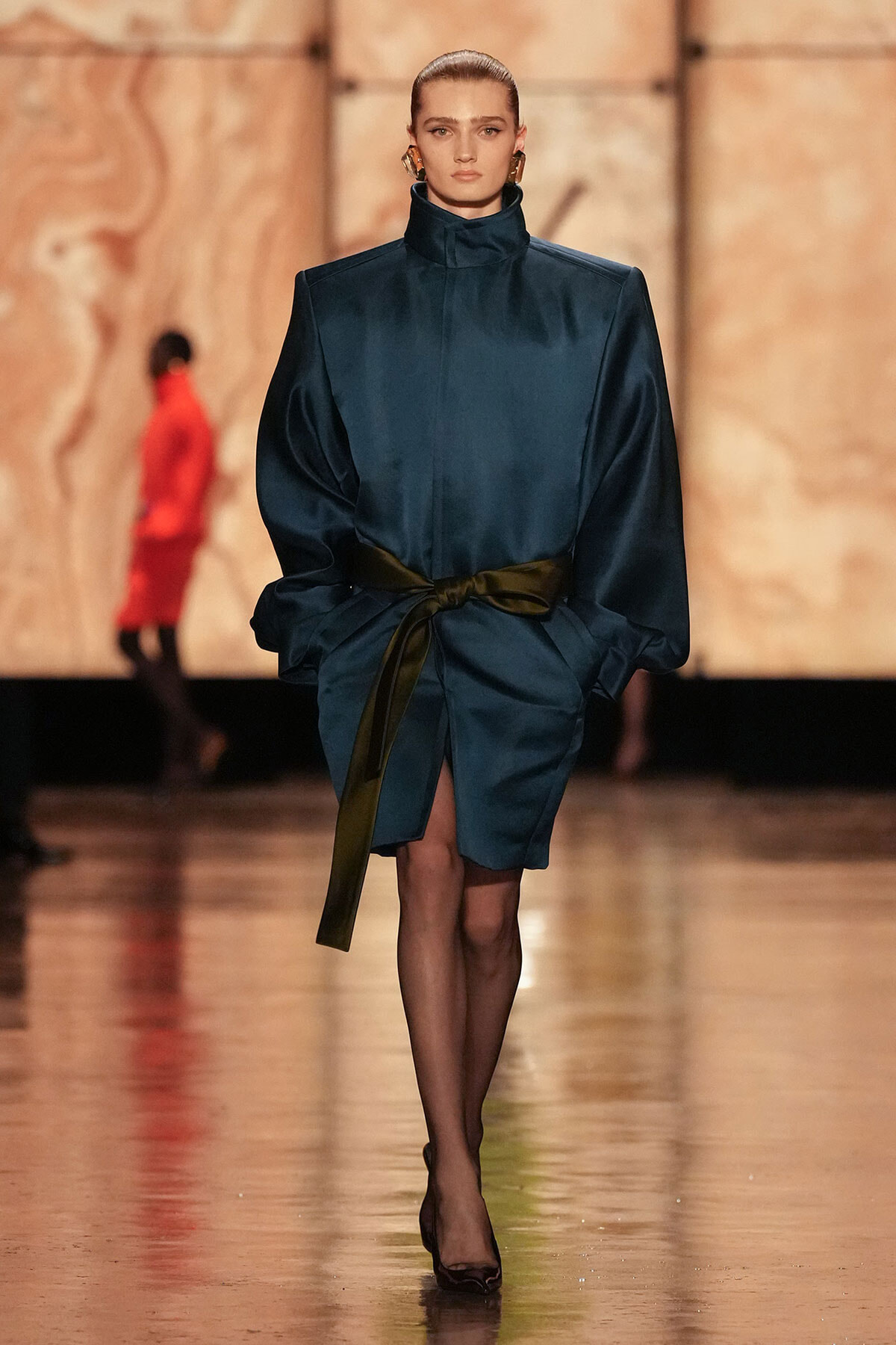 Model walks the runway in a teal satin dress with a high collar, voluminous sleeves, and a black sash tied at the waist, paired with sheer tights and black heels.