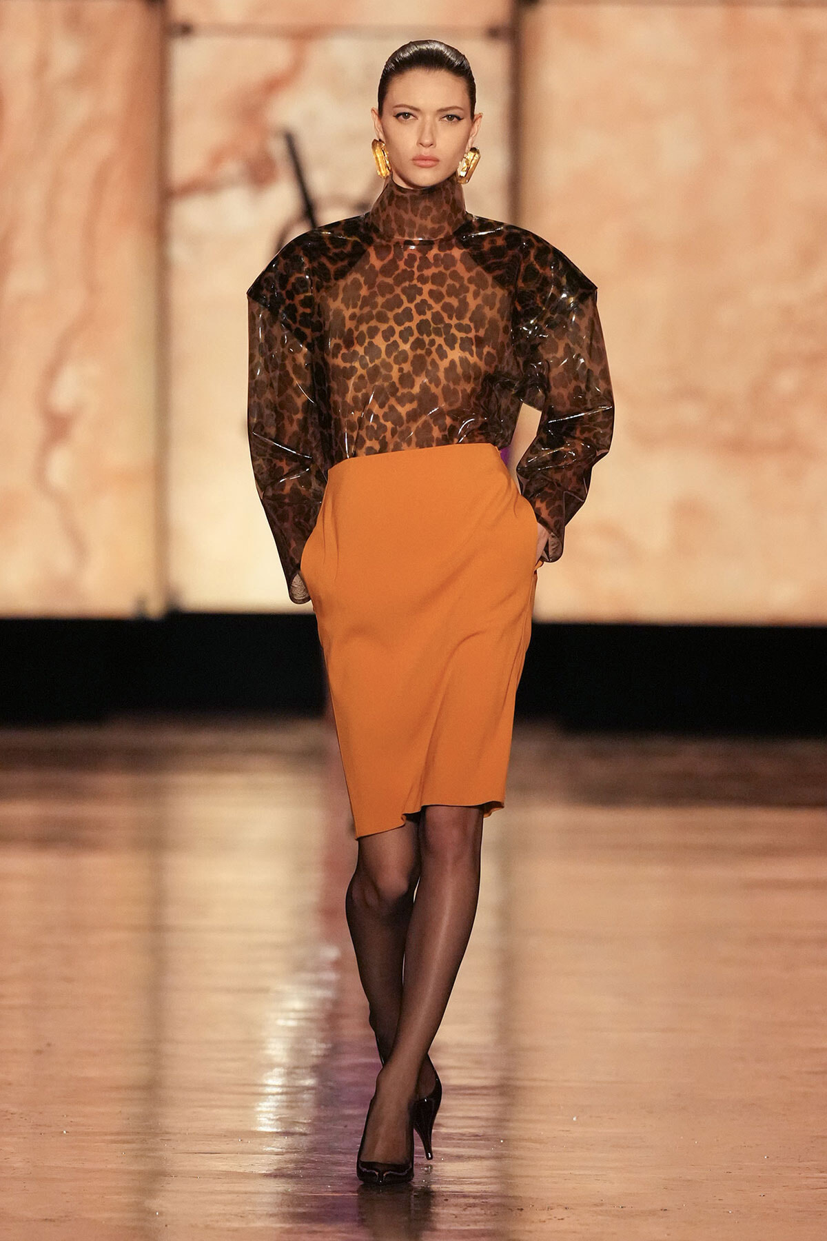 Model walking a fashion runway in a leopard-print sheer top tucked into an orange knee-length pencil skirt; black tights and black heels, bold gold earrings.
