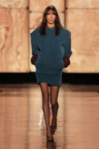 Model walking a fashion runway in a teal oversized dress, burgundy gloves, black tights, and beige heels against a warm backdrop.