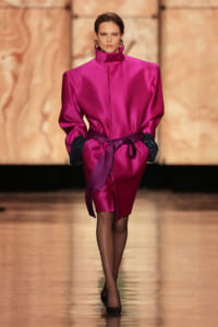 Model walking down runway in a bright pink satin coat with a tied belt and black gloves, posing in heels.