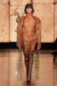 Model walking the runway in a sheer, brown marbled plastic dress with long sleeves and black tights.