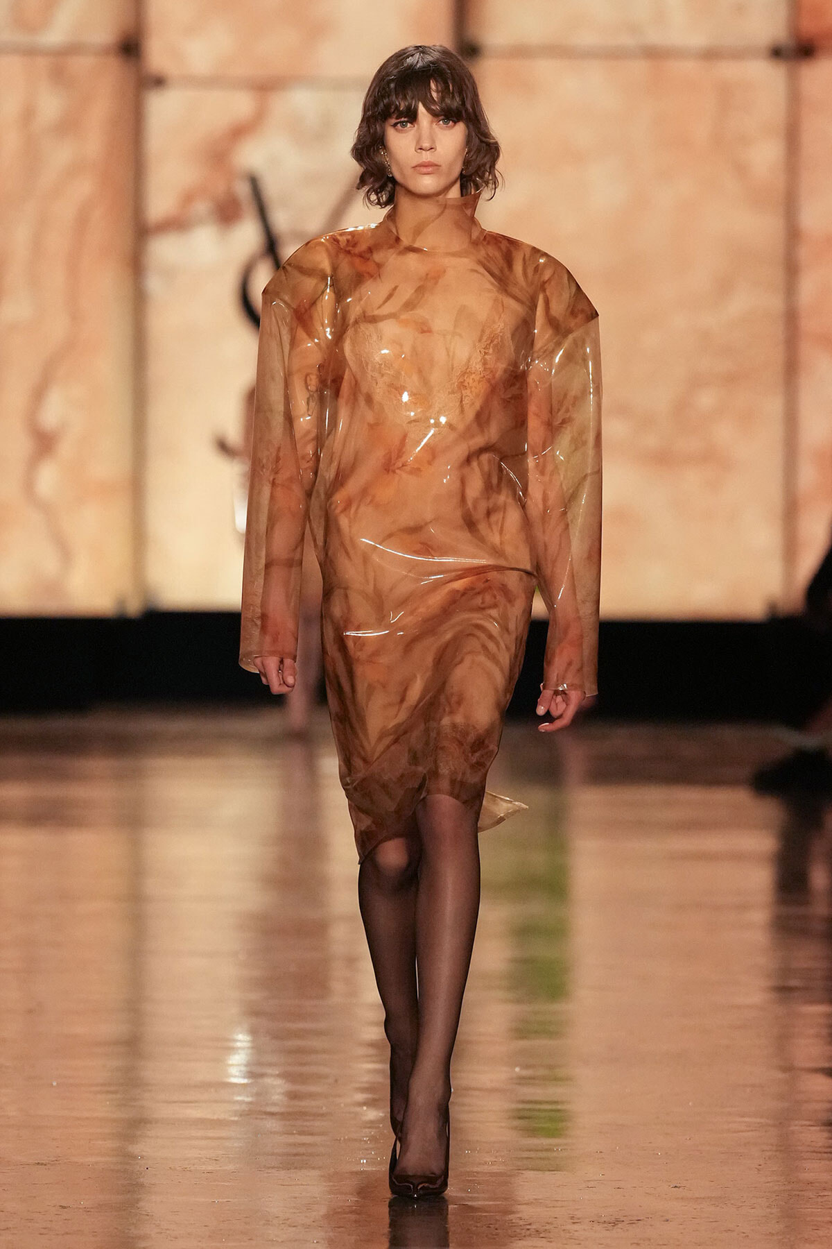 Model walking the runway in a sheer, brown marbled plastic dress with long sleeves and black tights.