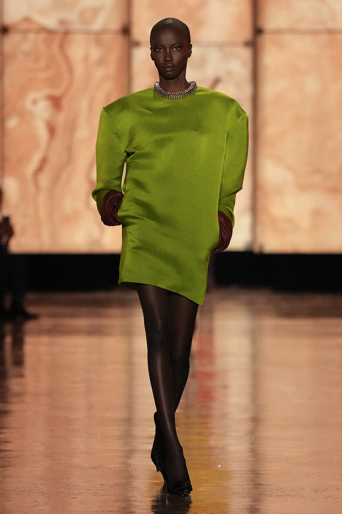 Model walking a lime-green, oversized dress with black tights and burgundy gloves on a runway, wearing a chunky necklace.