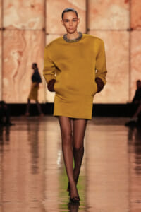 Model walking a fashion runway in a mustard-yellow mini dress with brown cuffed sleeves, hands in pockets, paired with sheer black tights, black heels, and a chunky beaded collar necklace.